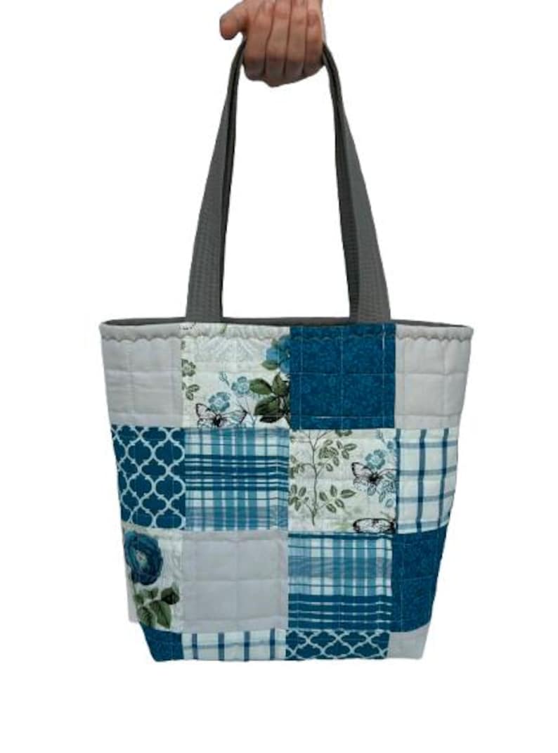 May include: A patchwork tote bag with a gray handle. The bag features a variety of blue and white patterned squares, including floral, plaid, and geometric designs. The bag is held up by a hand.
