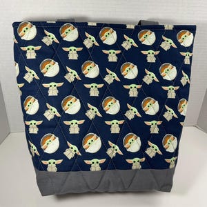 May include: A navy blue tote bag with a quilted pattern featuring a repeating design of a cartoon character. The bottom and handles are gray. The bag is designed to carry items.