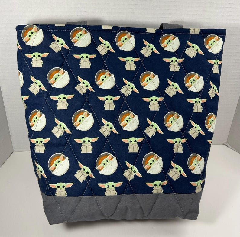 May include: A navy blue tote bag with a quilted pattern featuring a repeating design of a cartoon character. The bottom and handles are gray. The bag is designed to carry items.