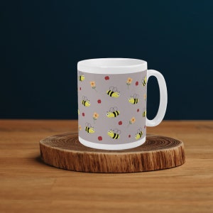 Puede incluir: Una taza de cerámica blanca con fondo gris y un patrón repetitivo de abejas amarillas y flores rojas y amarillas.