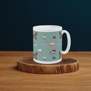 Puede incluir: Taza de cerámica blanca con un fondo verde azulado con dibujos de animales de granja, incluyendo caballos, cabras, conejos, cerdos y vacas. La taza tiene un asa blanca y está sobre un posavasos de madera.