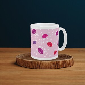 Puede incluir: Una taza de cerámica blanca con fondo rosa y un patrón de labios morados, rosas y rojos. La taza tiene un asa blanca.