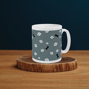 Puede incluir: Una taza de cerámica blanca con fondo gris y un patrón de huellas de patas blancas y formas de huesos negros.