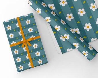 Pode incluir: Um papel de embrulho azul-esverdeado com um padrão de margaridas brancas e folhas amarelas. O papel de embrulho está enrolado e amarrado com uma fita laranja brilhante. Uma caixa de presente menor está embrulhada no mesmo papel e amarrada com a mesma fita.