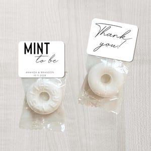 Op de afbeelding: Twee kleine, transparante cellofaan zakjes met elk een witte pepermunt. Eén zakje heeft een wit label met de tekst "MINT to be" en de namen "Amanda & Brandon" en de datum "10.11.2028". Het andere zakje heeft een wit label met de tekst "Thank you!"