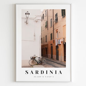 May include: A poster with a photograph of a narrow street in Sardinia, Italy. The street is lined with buildings in shades of peach and white. A bicycle is parked on the left side of the street. The poster has the text "SARDINIA" and the coordinates "40.1209° N 9.0129° E" printed at the bottom.