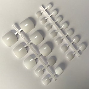 May include: Multiple sets of white, square-shaped press-on toenails are displayed on a white surface. The nails have a glossy finish, suggesting they are ready for use. The image is well-lit, emphasizing the smooth texture of the artificial nails.