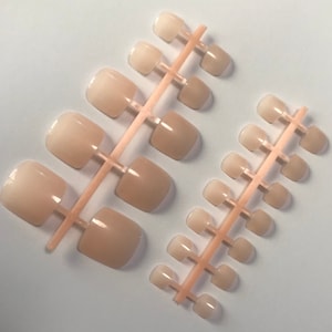 May include: Two sets of nude-coloured press-on toenails arranged on light pink plastic frames. The set on the left features larger, square-shaped nails, while the set on the right contains smaller, square-shaped nails in a matching glossy finish.