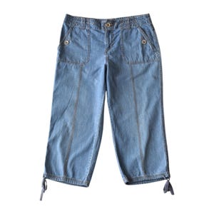 May include: Light blue denim capri pants with two front pockets and two back pockets. The pants have a button closure and decorative stitching. The hemline features adjustable ties. The pants are made of a lightweight denim material.