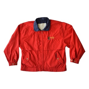 May include: A red jacket with a navy blue collar and cuffs. The jacket has a zipper closure, two front pockets, and buttoned cuffs. The jacket has the text "Waterfall Alaska" embroidered on the chest.