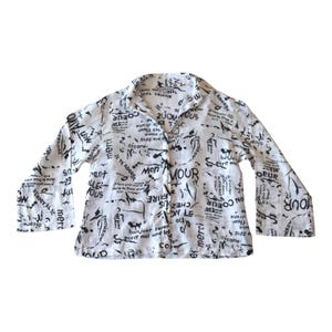 May include: White button-down shirt with long sleeves. The shirt is covered in black text and abstract shapes. The collar and cuffs are visible, and the shirt is unbuttoned.