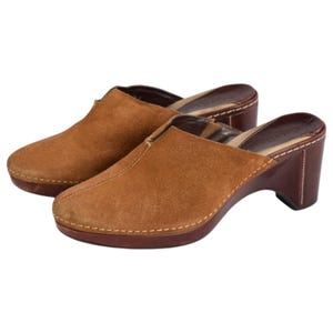 May include: A pair of brown suede clogs with a slip-on design. The shoes have a rounded toe, a dark brown sole, and a stacked heel. The stitching is visible around the edges of the sole and heel.