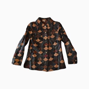 Puede incluir: Una camisa de manga larga con botones, confeccionada con parches de cuero multicolor dispuestos en un patrón repetitivo de escamas. La prenda presenta un cuello en punta, dos bolsillos en el pecho y botones metálicos en la parte frontal.