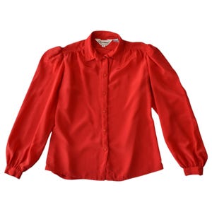 Può includere: Una camicia rossa a maniche lunghe con abbottonatura frontale, colletto classico, spalle a sbuffo arricciate e polsini. Il capo presenta una fila verticale centrale di bottoni rossi abbinati e un'etichetta bianca sul collo.