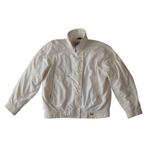 May include: Cream-coloured jacket with a stand-up collar, zip, and button closure. The jacket has long sleeves, pockets, and a ribbed hem. The material appears lightweight and slightly textured. The jacket is a neutral colour.