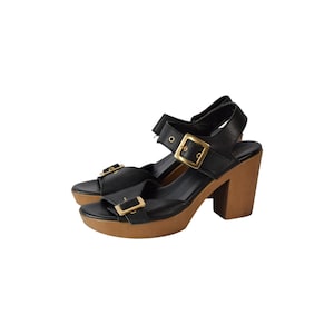 Op de afbeelding: Een paar zwarte leren sandalen met een dikke houten plateauzool en een stevige houten blokhak. Elke sandaal heeft twee verstelbare bandjes over de wreef en bij de enkel, vastgezet met grote, vierkante, goudkleurige metalen gespen.