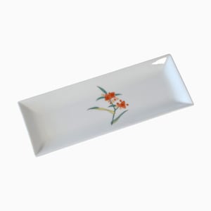 Può includere: Un vassoio rettangolare in ceramica bianca con un'illustrazione botanica centrale di fiori arancioni con foglie verdi e sottili steli gialli. Il vassoio presenta bordi leggermente rialzati e una finitura liscia e lucida sulla superficie.