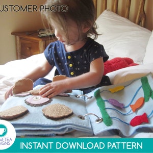 May include: A child plays with a felt quiet book featuring a blue ocean scene with fish and a clam with a pearl inside. The book is made of felt and has a blue background. The book is on a white bed.
