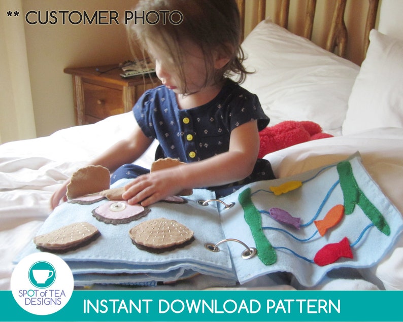 May include: A child plays with a felt quiet book featuring a blue ocean scene with fish and a clam with a pearl inside. The book is made of felt and has a blue background. The book is on a white bed.
