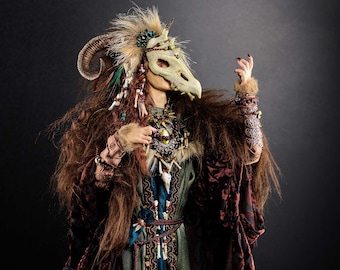 May include: A fantasy figure wearing a long, burgundy and green robe with a bone and bead embellishment. The figure is wearing a bone mask with a long snout and horns. The figure is standing on a wooden base.