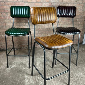 May include: Three bar stools with black metal frames and ribbed, padded seats and backs. The stools have seats and backs in green, tan, and dark brown. The stools are set against a brick wall.