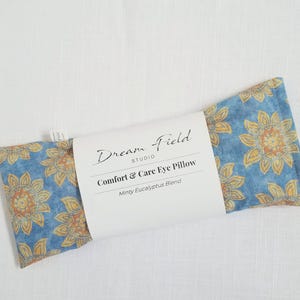 May include: A blue and yellow floral Comfort & Care Eye Pillow from Dream Field Studio. The pillow has a floral pattern and the text "Comfort & Care Eye Pillow" and "Minty Eucalyptus Blend" on the label.