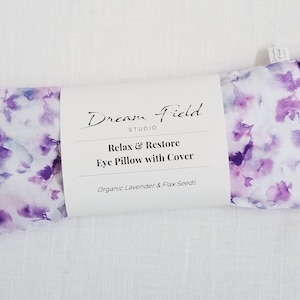 Op de afbeelding: Een oogkussen met een bloemenpatroon in paars en wit. Het kussen is verpakt met een wit label met de tekst "Dream Field Studio" en "Relax & Restore Eye Pillow with Cover". Het bevat biologische lavendel en lijnzaad.