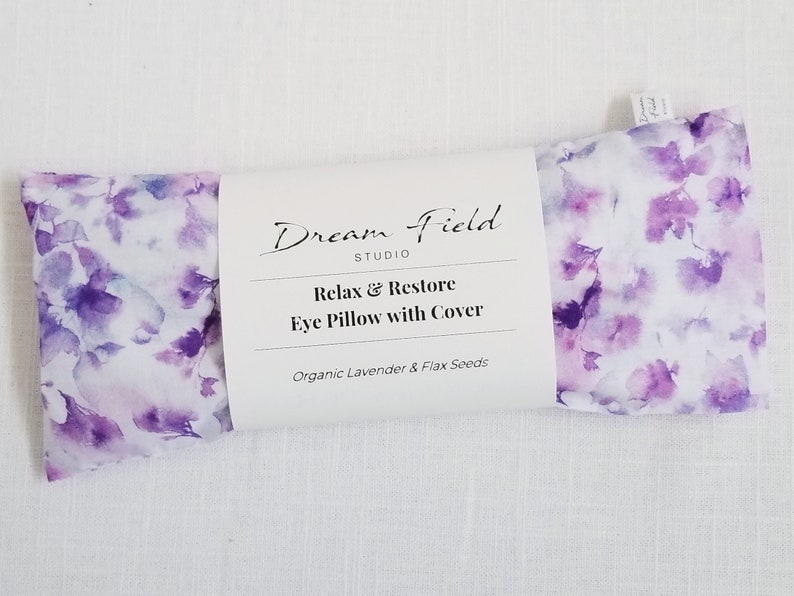 Op de afbeelding: Een oogkussen met een bloemenpatroon in paars en wit. Het kussen is verpakt met een wit label met de tekst "Dream Field Studio" en "Relax & Restore Eye Pillow with Cover". Het bevat biologische lavendel en lijnzaad.
