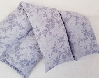 May include: A lavender-colored, rectangular, floral-patterned heating pad. The fabric features a repeating design of delicate flowers and leaves in a darker shade of lavender. The pad is folded over on itself.