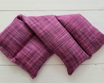 May include: A rectangular heating pad featuring a textured fabric in shades of magenta, purple, and black. The fabric has a cross-hatched, woven appearance with visible white and dark threads throughout, creating a multi-tonal, deep pink aesthetic.
