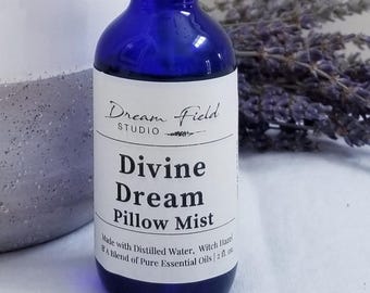 May include: A cobalt blue glass bottle with a white label featuring the text "Dream Field Studio Divine Dream Pillow Mist." The label also states the product is made with distilled water, witch hazel, and essential oils. The bottle contains 2 fl oz.