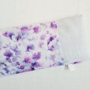 Op de afbeelding: Een rechthoekig stoffen item met een bloemenpatroon in paarse en witte tinten. Het ontwerp lijkt op aquarelbloemen. Het item is gedeeltelijk open en onthult een effen wit interieur.