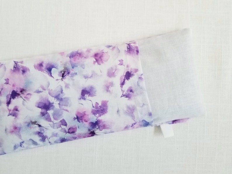 Op de afbeelding: Een rechthoekig stoffen item met een bloemenpatroon in paarse en witte tinten. Het ontwerp lijkt op aquarelbloemen. Het item is gedeeltelijk open en onthult een effen wit interieur.