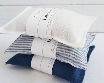 May include: A stack of three lavender sachets in white, blue striped, and solid navy blue fabrics. Each sachet is wrapped with a white label that reads "Lavender" and "Organic Lavender Buds". The sachets are stacked on top of each other.