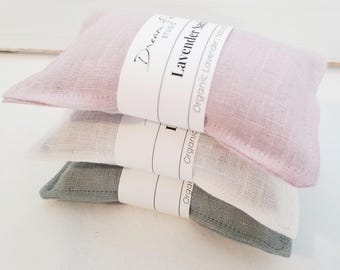 May include: Three rectangular fabric sachets stacked vertically, colored light pink, white, and sage green. Each sachet features a white paper band with the printed text Dream Studio, Lavender Sachet, and Organic Lavender.