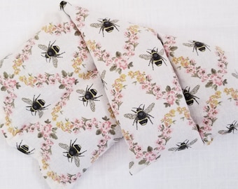 May include: Three rectangular fabric items with a floral and bee pattern. The pattern features black and yellow bees and pink and yellow flowers on a white background. The items are arranged on a white surface.