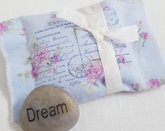 May include: A light blue fabric pouch with a floral design and a white ribbon, next to a smooth, gray stone with the word "Dream" printed on it. The pouch has a vintage postcard print with pink roses.