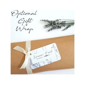 May include: A wrapped gift in brown paper, tied with a cream ribbon and a tag that reads "Dream Field Studio." The words "Optional Gift Wrap" are written in cursive above the gift. A sprig of lavender is placed on the white background.