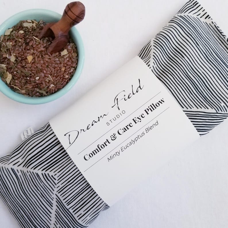 May include: An eye pillow with a black and white striped pattern, labeled "Comfort & Care Eye Pillow" and "Minty Eucalyptus Blend." A small blue bowl with a wooden scoop and a mix of seeds and herbs is next to the pillow. The brand name "Dream Field Studio" is also visible.