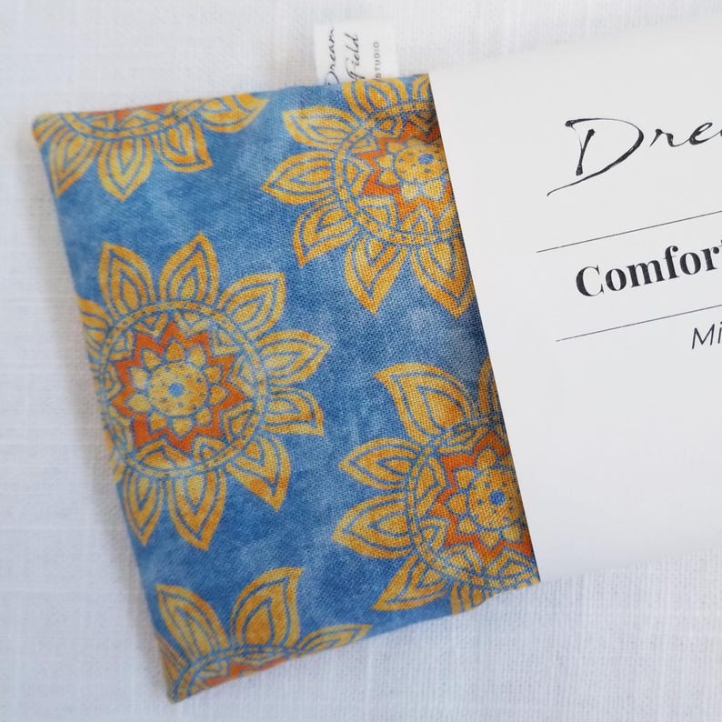 May include: A blue fabric item with a repeating pattern of yellow and orange floral designs. The item is partially tucked into a white card with the text "Dream Field Studio" and "Comfort".