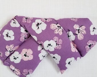May include: A lavender-colored, rectangular eye pillow with a floral pattern. The pattern features white and pink flowers with black centers. The pillow is folded and has a stitched edge.