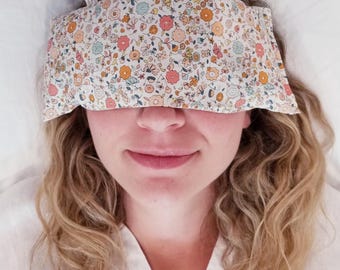 May include: A rectangular eye pillow featuring a floral pattern with small pink, orange, yellow, and light blue flowers on a white background. The fabric mask covers the upper portion of the face, resting across the eyes and bridge of the nose.