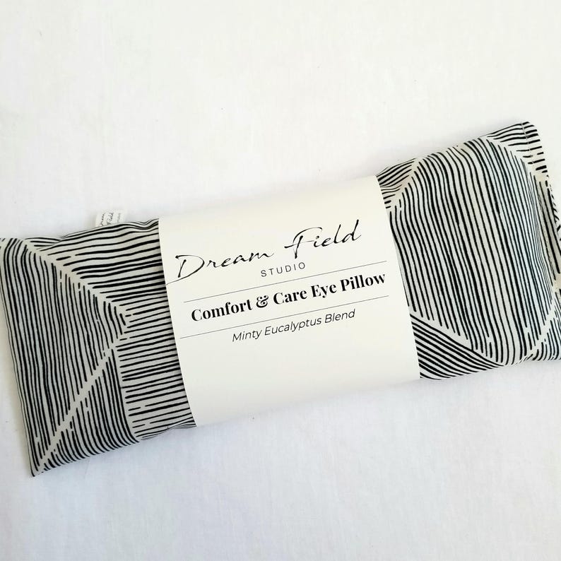 May include: A rectangular eye pillow with a black and white striped pattern. The pillow has a white label that reads "Dream Field Studio, Comfort & Care Eye Pillow, Minty Eucalyptus Blend."