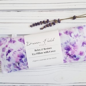 Op de afbeelding: Een lavendelgeurend oogkussen met een aquarel bloemenpatroon in paarse en witte tinten. Het kussen is voorzien van het label "Dream Field Studio" en "Relax & Restore Eye Pillow with Cover". Een takje lavendel ligt erbovenop.