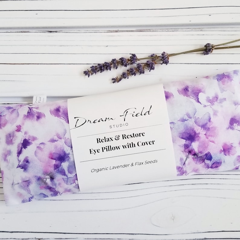 Op de afbeelding: Een lavendelgeurend oogkussen met een aquarel bloemenpatroon in paarse en witte tinten. Het kussen is voorzien van het label "Dream Field Studio" en "Relax & Restore Eye Pillow with Cover". Een takje lavendel ligt erbovenop.