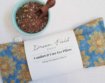 May include: A rectangular eye pillow with a blue fabric featuring a yellow sunflower pattern. A white paper band wraps around the center with the text Dream Field STUDIO Comfort & Care Eye Pillow Minty Eucalyptus Blend. A light blue bowl contains flax seeds and a small wooden scoop.