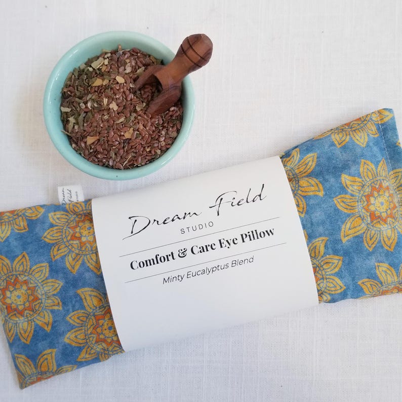 May include: A blue eye pillow with a yellow floral pattern, labeled "Dream Field Studio Comfort & Care Eye Pillow." A small bowl of herbs and a wooden scoop are next to the pillow. The pillow is filled with a minty eucalyptus blend.