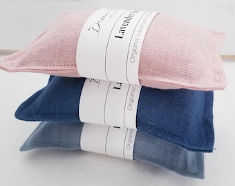 May include: A stack of three rectangular fabric sachets in shades of dusty pink, navy blue, and slate blue. Each sachet is wrapped in a white paper band featuring the text Dream Lavender Sachet and Organic Lavender. The fabric has a visible weave.