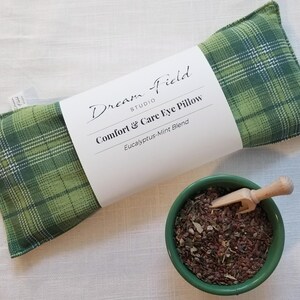 May include: A green plaid eye pillow with a white label that reads "Dream Field Studio Comfort & Care Eye Pillow Eucalyptus-Mint Blend." A small green bowl contains dried herbs and a wooden scoop.