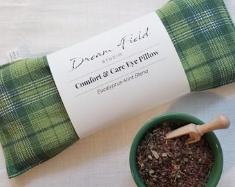 May include: A rectangular green plaid eye pillow with a white paper band labeled Dream Field Studio Comfort & Care Eye Pillow Eucalyptus-Mint Blend, accompanied by a small green bowl filled with brown seeds and a tiny wooden scoop.